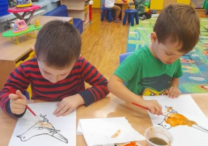 Dwoje dzieci siedzi przy stoliku w sali i maluje farbami